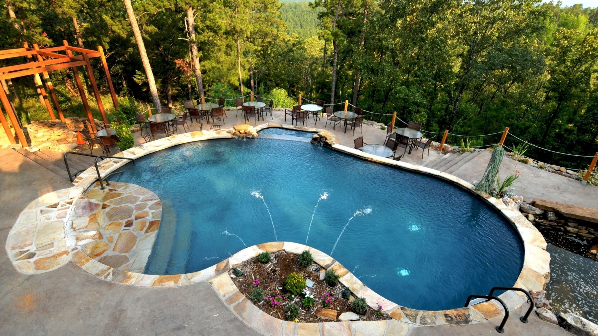 Luxury curved pool with fountains surrounded by lounge chairs and forest trees.