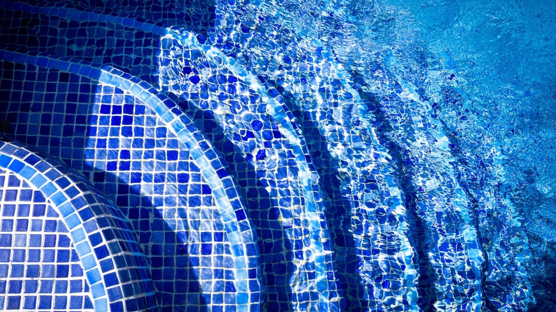 Underwater view of swimming pool tiles with rippling water patterns and light reflections.