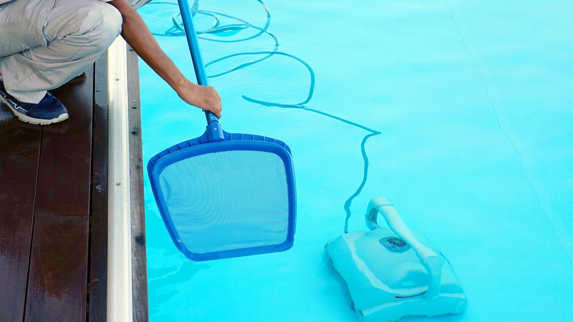 Person cleaning swimming pool with net skimmer and maintenance equipment.