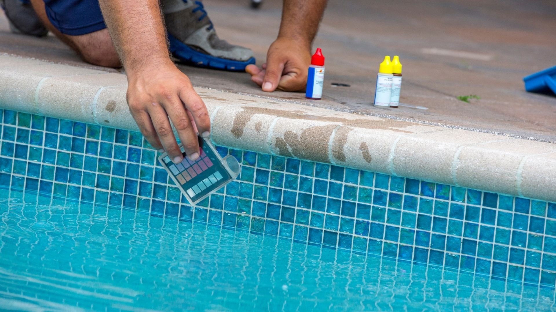 Worker testing pool water chemistry with color comparison kit and chemical bottles nearby.