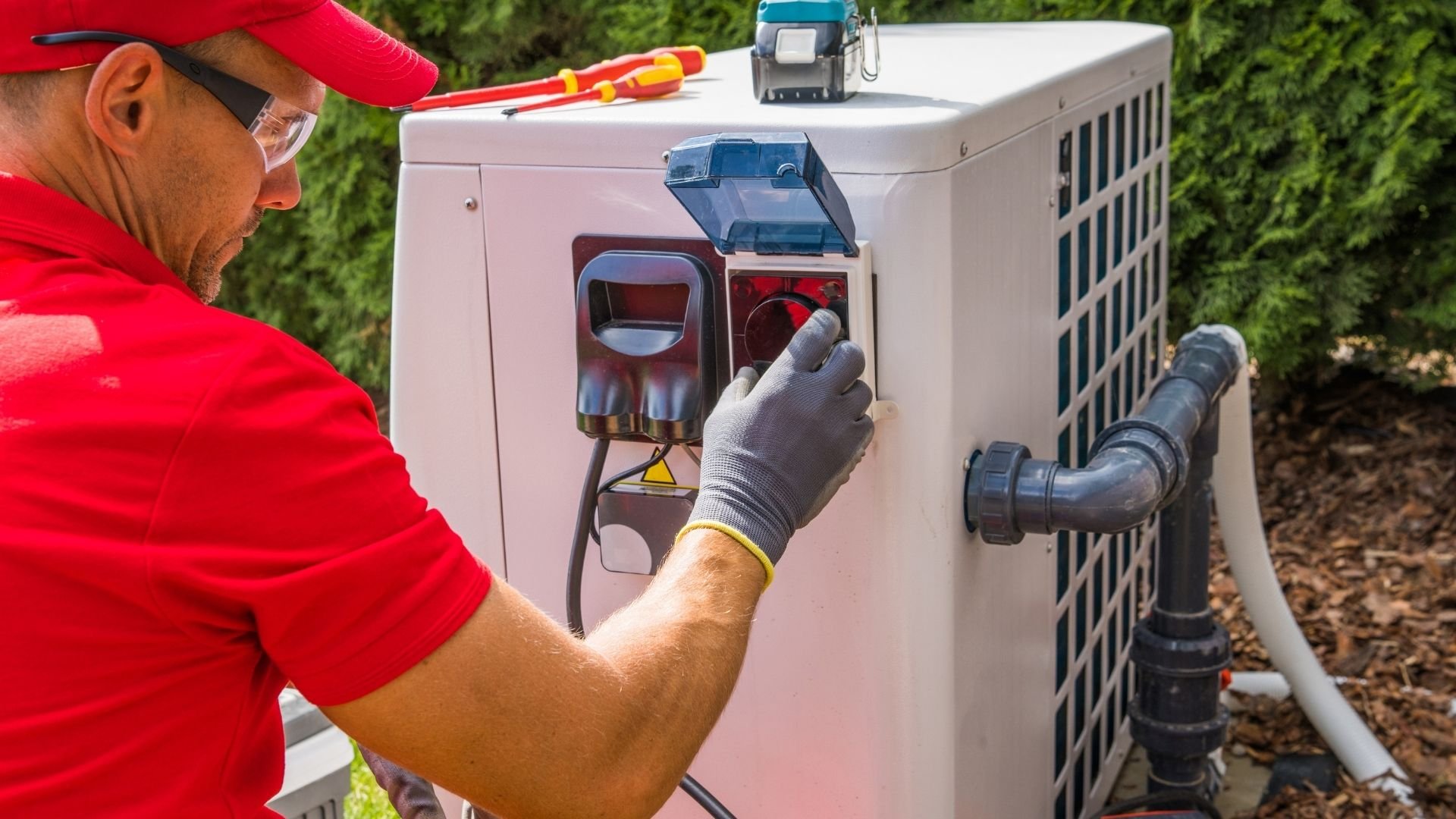 Technician in red uniform servicing outdoor HVAC air conditioning unit with tools