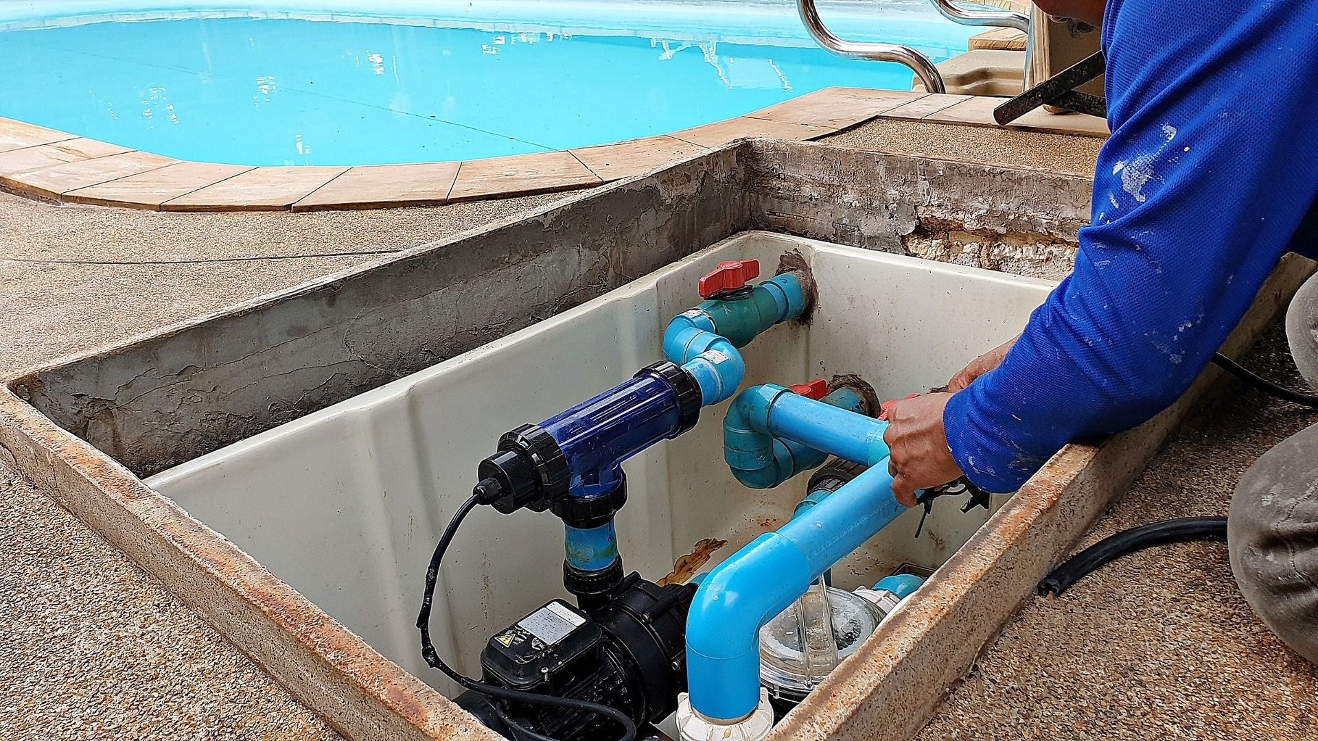 Worker in blue jacket maintains pool pump and filtration equipment beside turquoise swimming pool