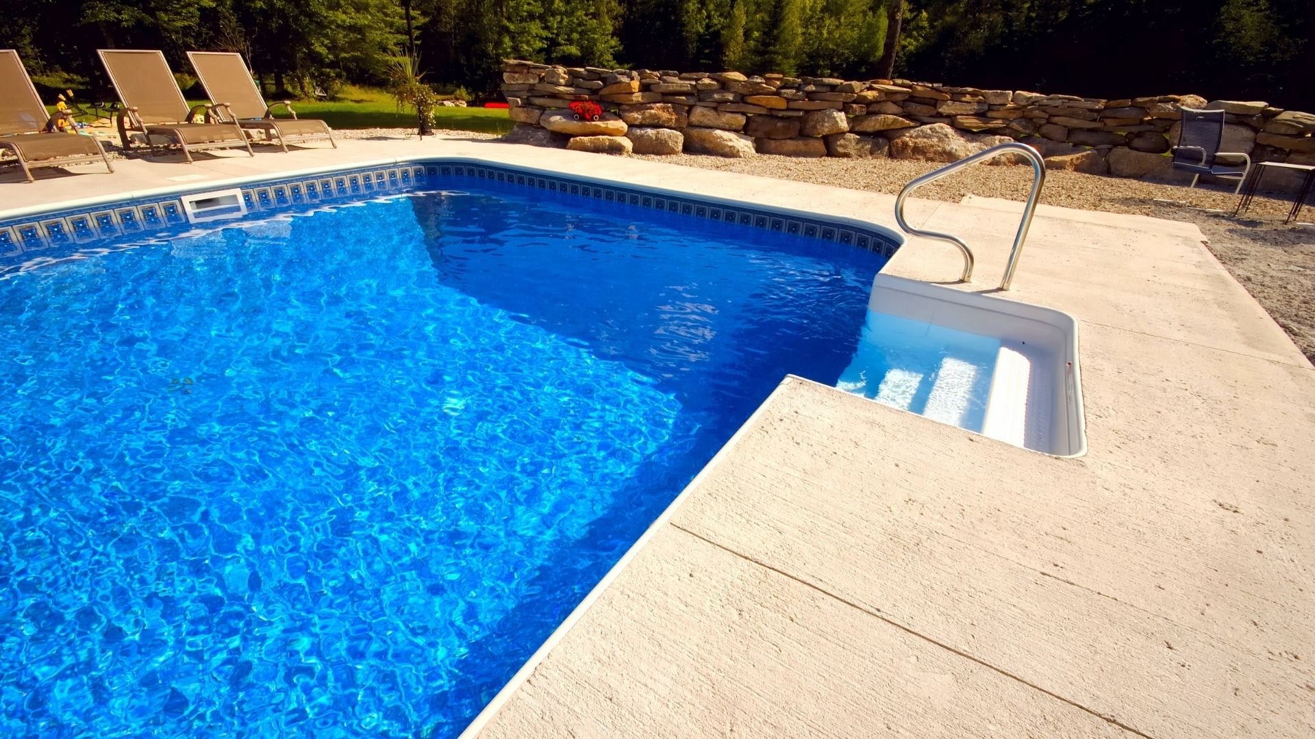 Sparkling blue backyard swimming pool with deck chairs, stone wall, and evergreen trees