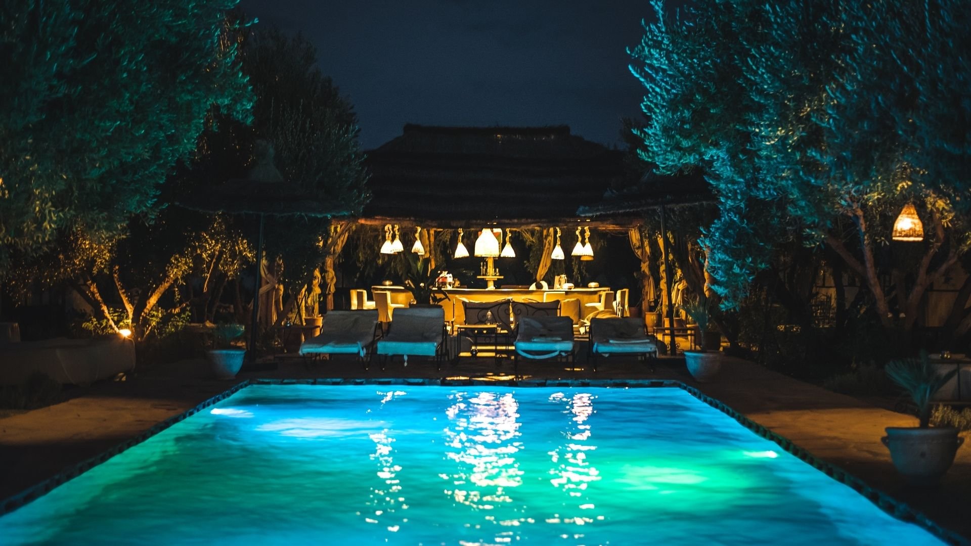 Illuminated swimming pool at night with dining pavilion, lounge chairs, and glowing trees