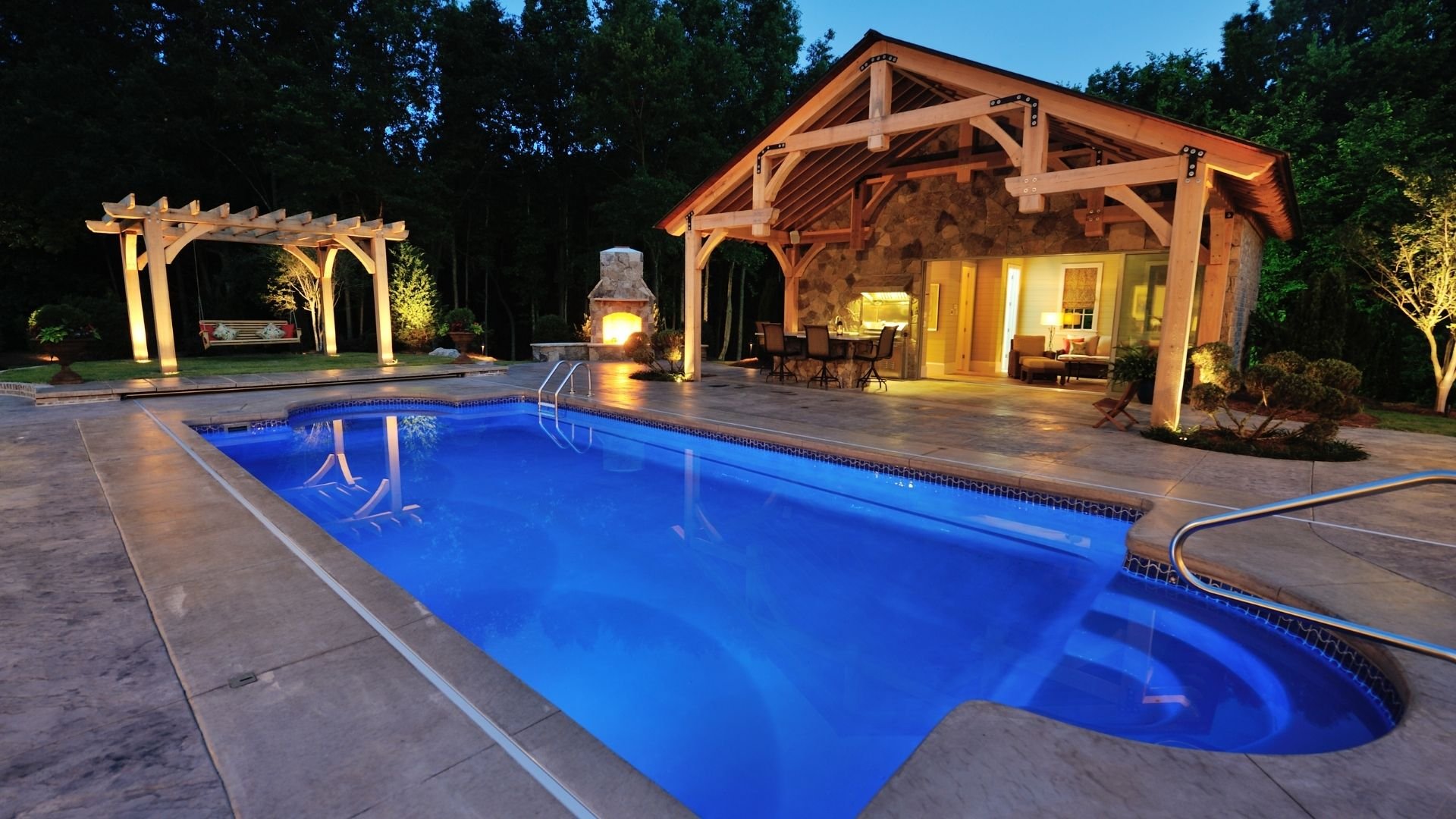 Illuminated backyard pool with modern wooden pavilion and fireplace at dusk