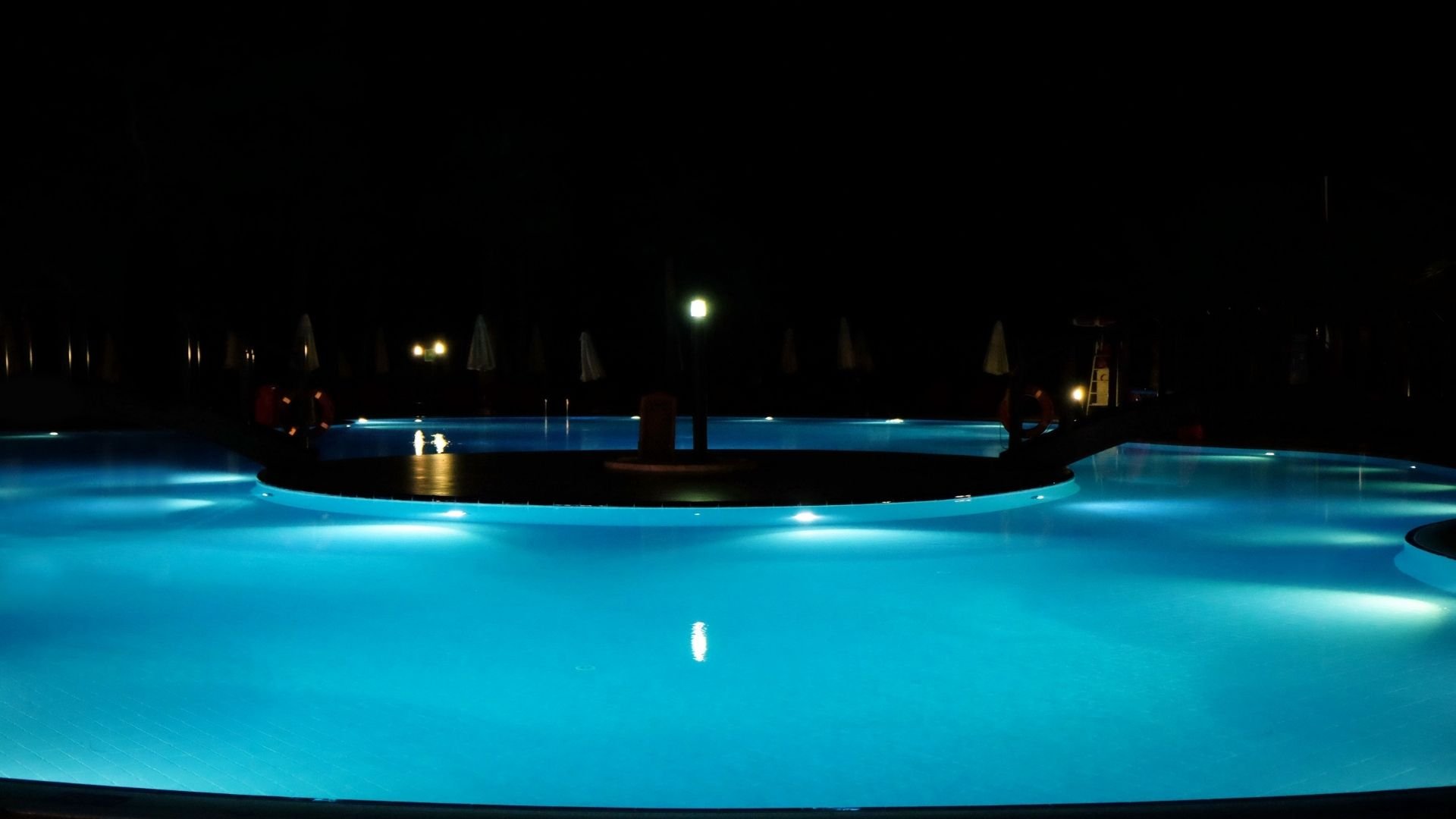 Illuminated resort pool at night with glowing turquoise water and umbrellas