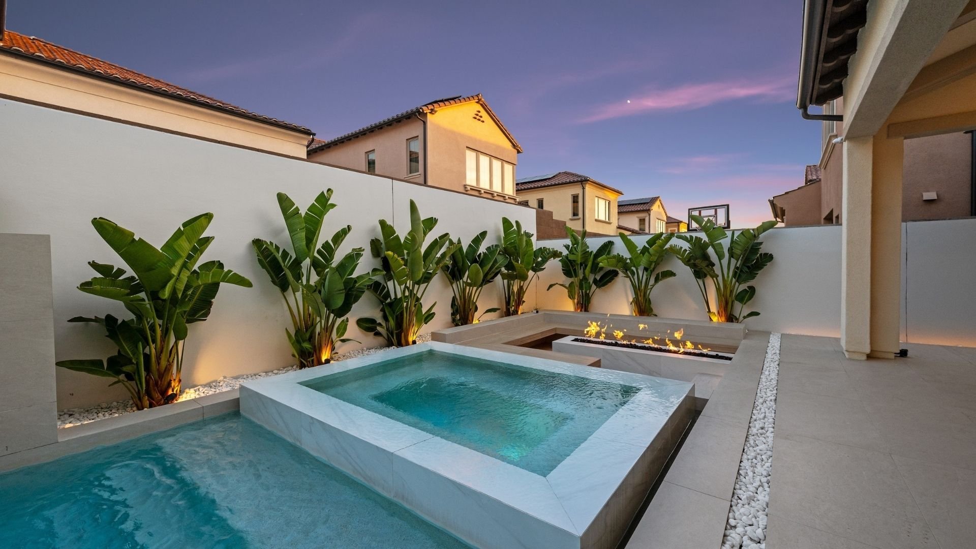Luxury backyard with illuminated pools, hot tub, fire pit, tropical plants, at twilight
