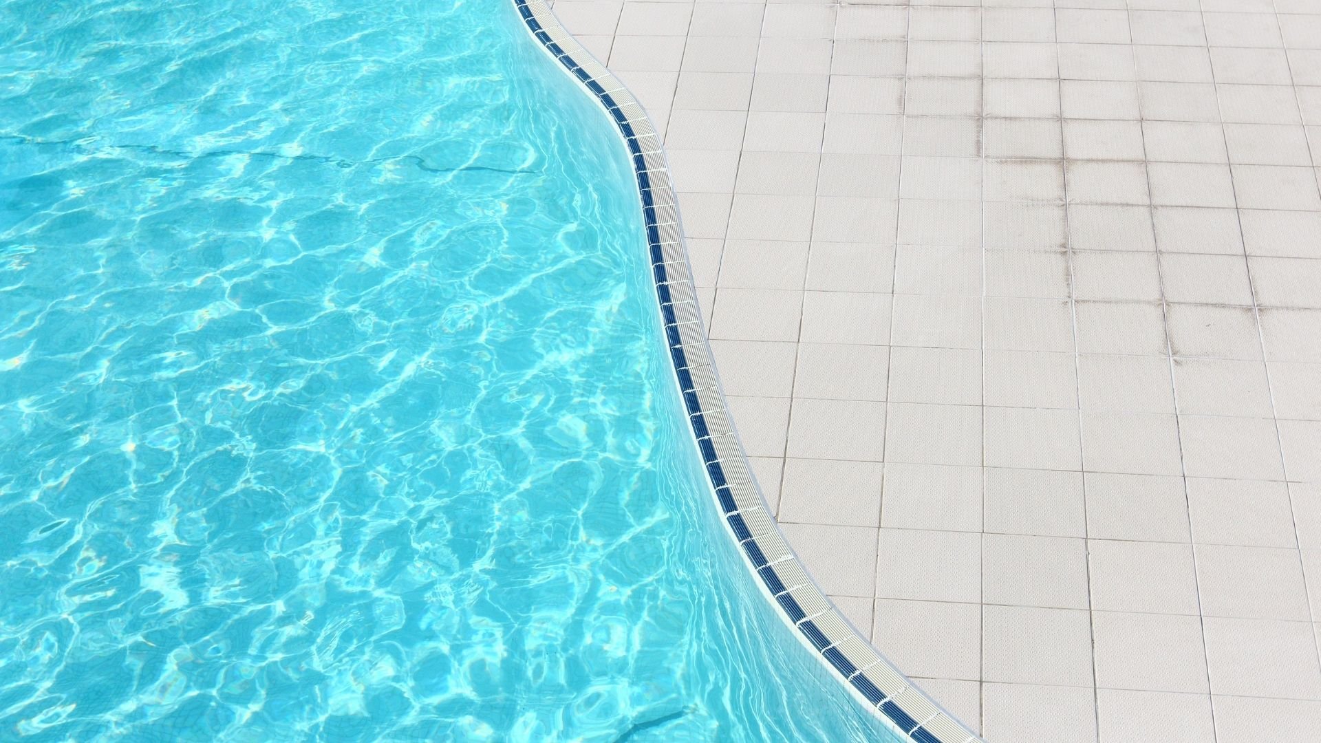 Overhead view of bright turquoise swimming pool water next to white tiled pool deck