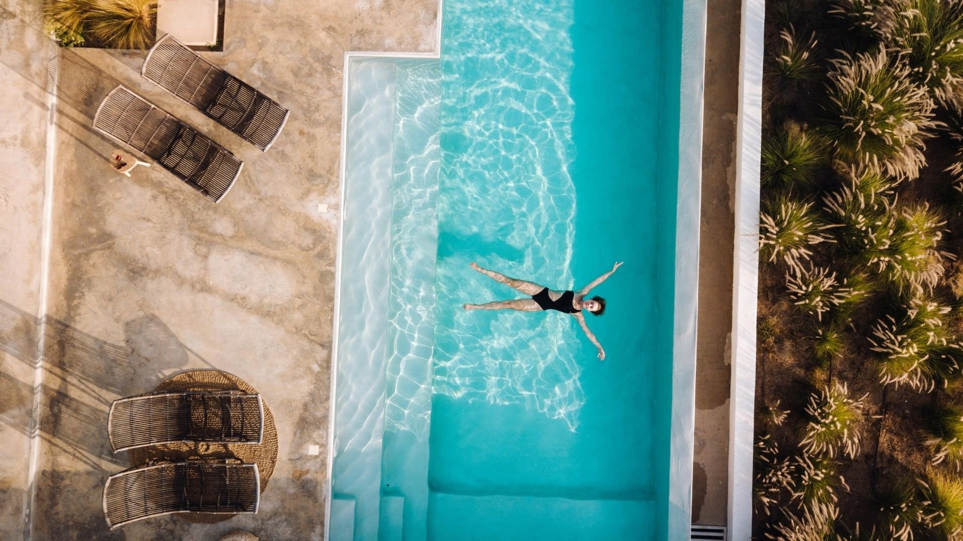 Aerial view of person swimming in bright turquoise pool surrounded by loungers and tropical plants