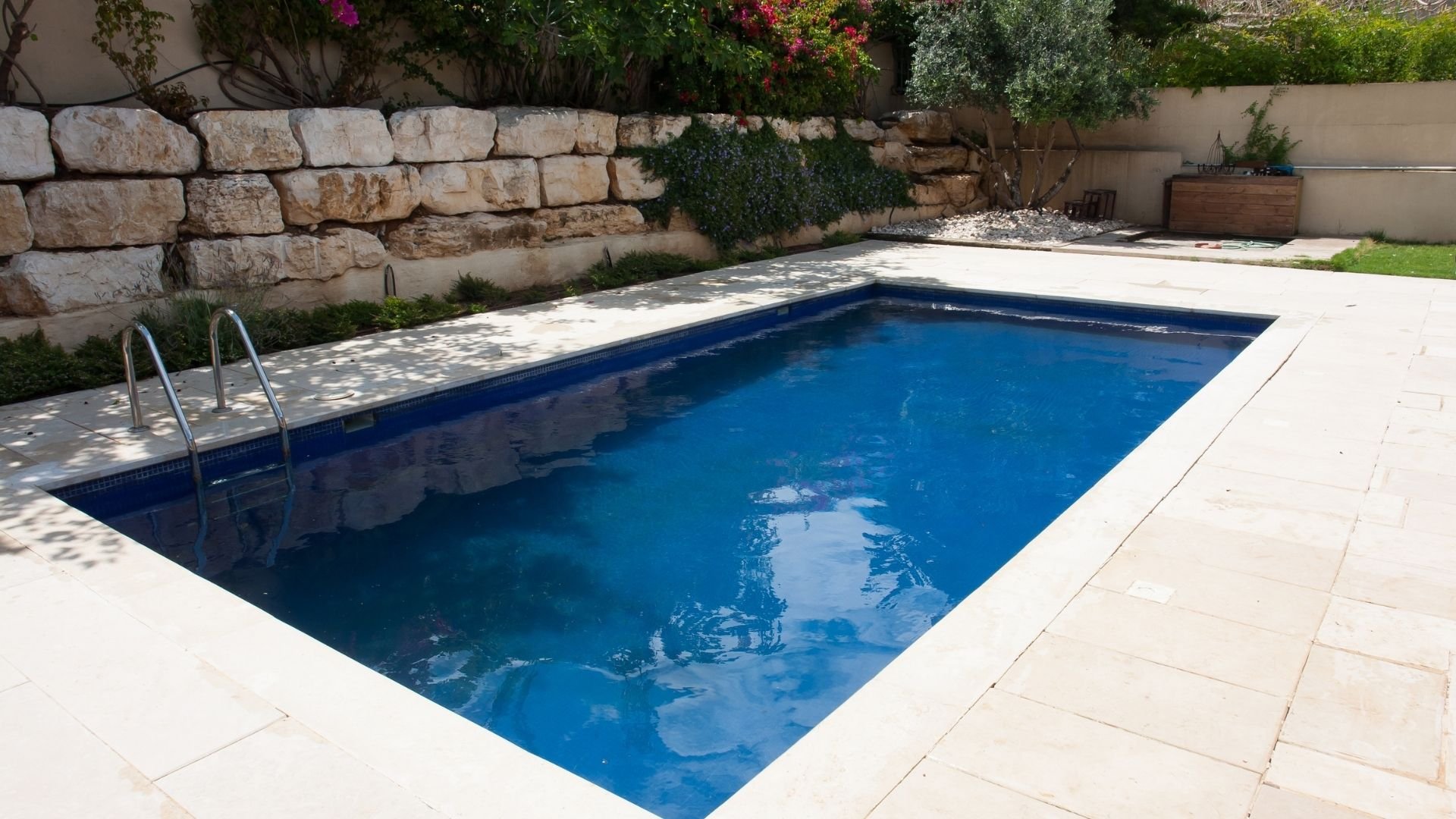 Rectangular swimming pool with blue water, metal ladder, surrounded by stone walls and flowering plants